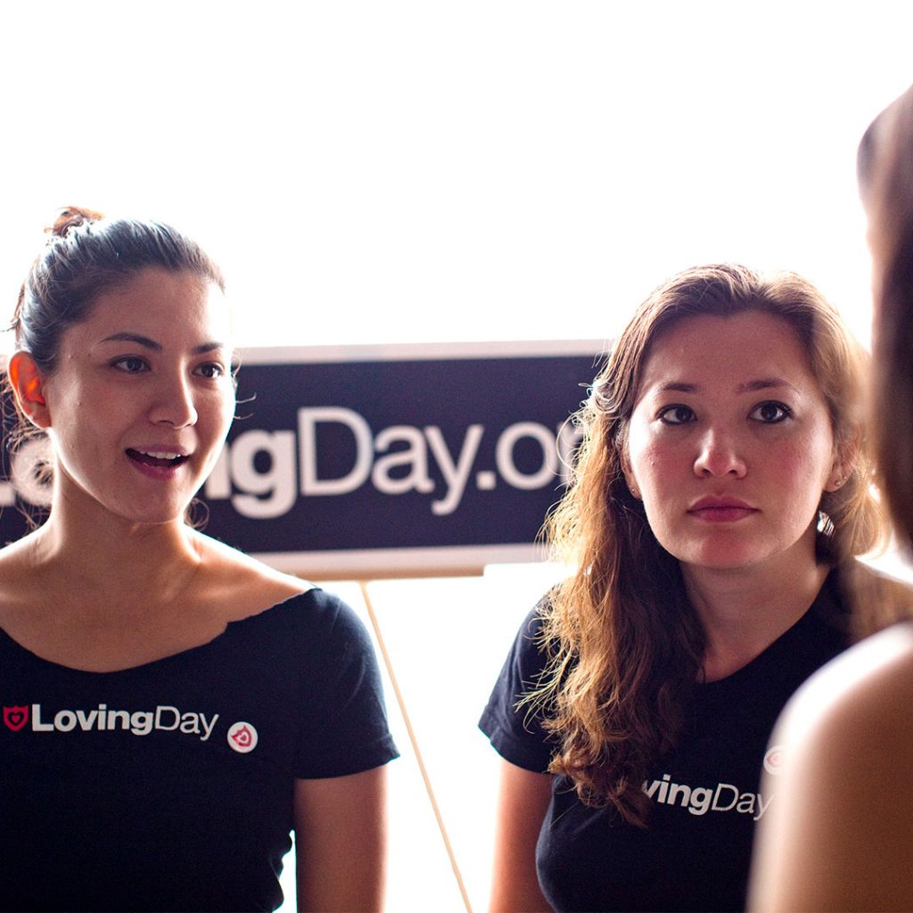 Two women volunteering and wearing Loving Day t-shirts who are standing behind a Loving Day sign and speaking to a guest at Loving Day NYC 2010.