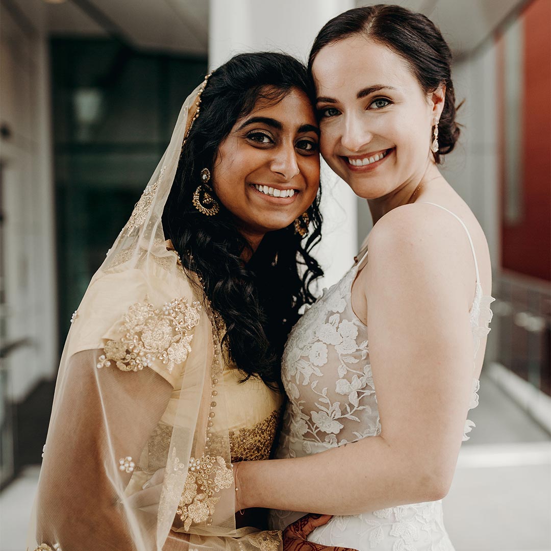 A wedding photo of two women outdoors, facing forward, smiling, and holding hands.